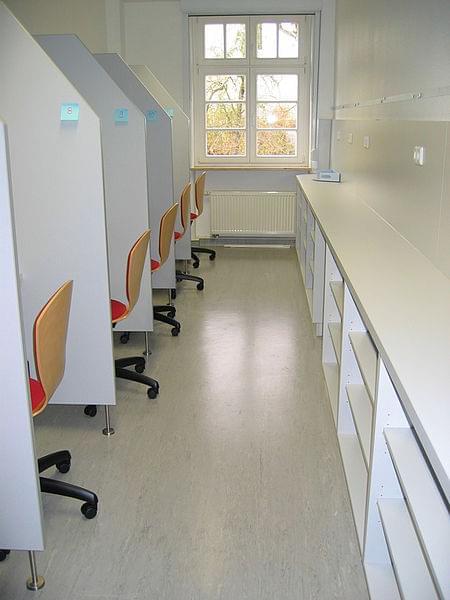 Ein schmaler Raum, links eine Reihe Testplätze mit Trennwänden, rechts ein langes Regal